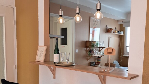 A dining area with a wooden bar counter, two black upholstered chairs, hanging light fixtures, and a mirror above the counter reflecting the room. - Large, cozy, quiet family apartment in the center of town, with view of the cathedral (Strasbourg)