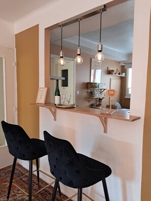 Een eetgedeelte met een houten bar, twee zwarte fauteuils, hanglampen en een spiegel boven de bar die de ruimte weerspiegelt.