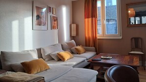 A modern living room with a sofa, a coffee table, and a chair. There are framed pictures on the wall, a window with curtains, and a lamp on the table. - Large, cozy, quiet family apartment in the center of town, with view of the cathedral (Strasbourg)