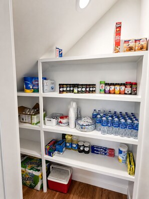 Uma despensa bem organizada com diversos alimentos, incluindo temperos, água engarrafada e produtos de limpeza.