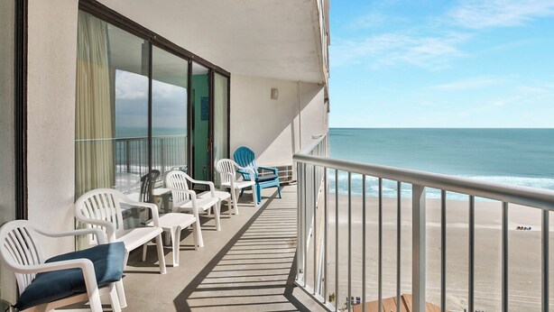 Ein Balkon mit weißen Plastikstühlen und einem Tisch mit Blick auf einen Strand und einen klaren Himmel.