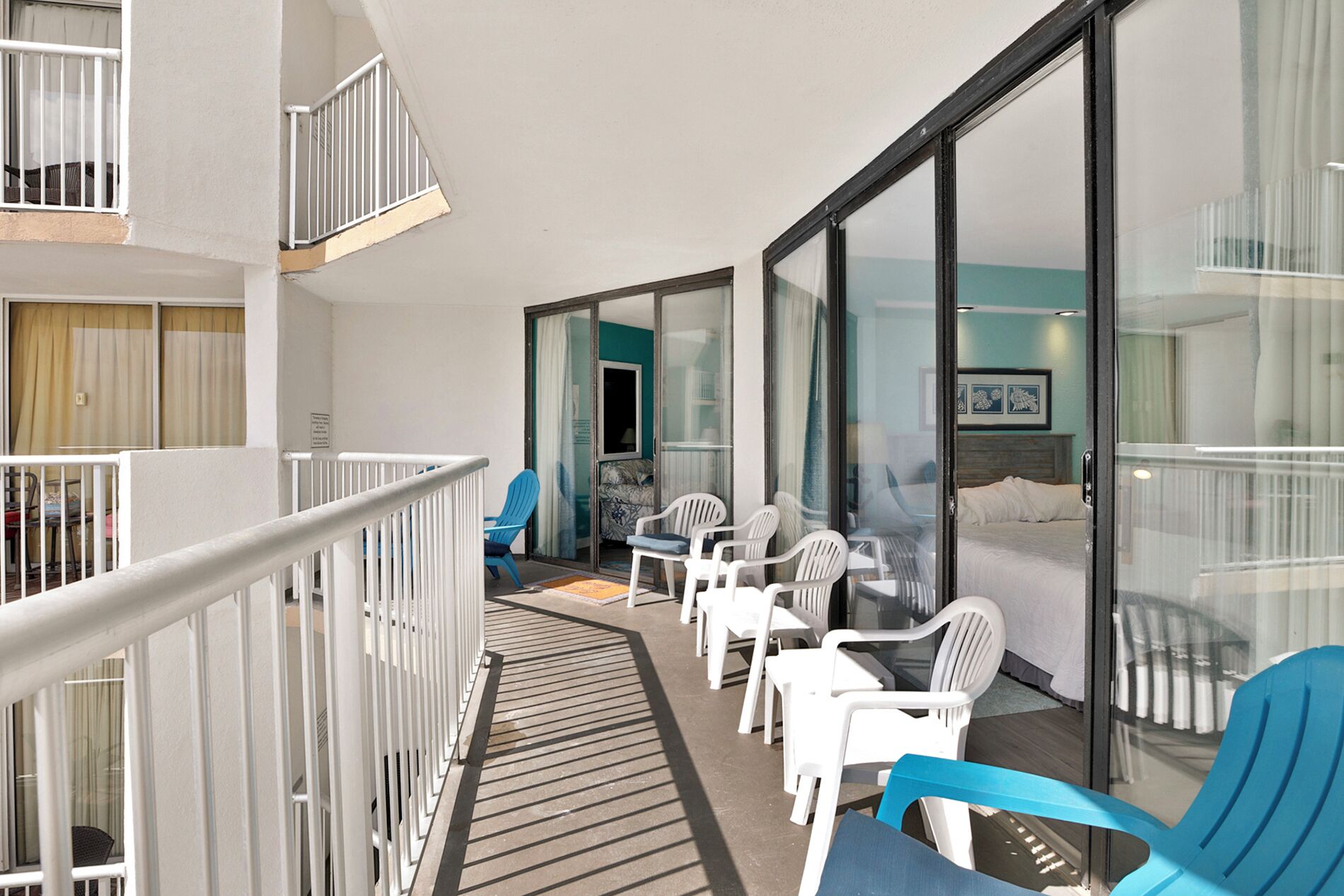 A balcony with white plastic chairs and a blue chair, a bed with white bedding, and a glass door leading to a room.