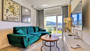 Un salon moderne avec un canapé vert, une table basse ronde en bois et une vue sur un balcon aménagé avec du mobilier d’extérieur.