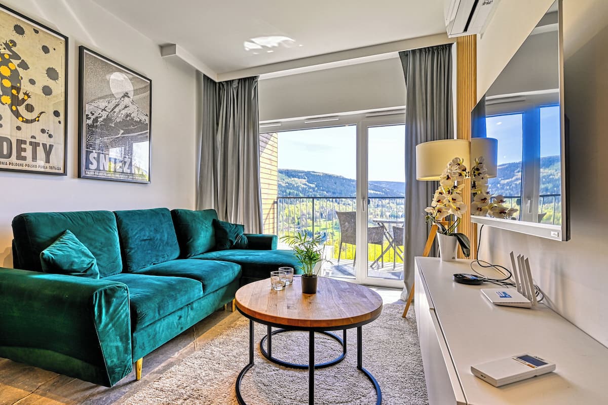 Un salon moderne avec un canapé vert, une table basse ronde en bois et une vue sur un balcon aménagé avec du mobilier d’extérieur.