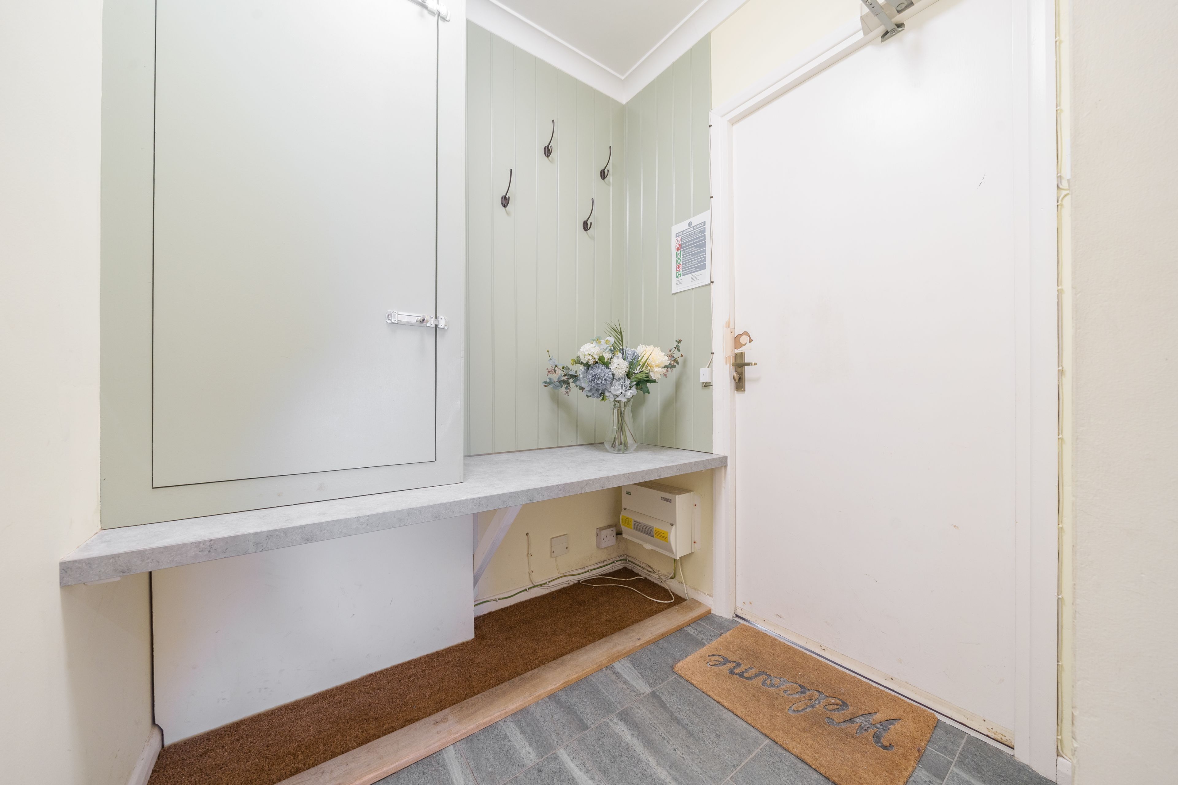 A hallway with a white door, a small shelf with a vase of flowers, and a doormat with the word "welcome".