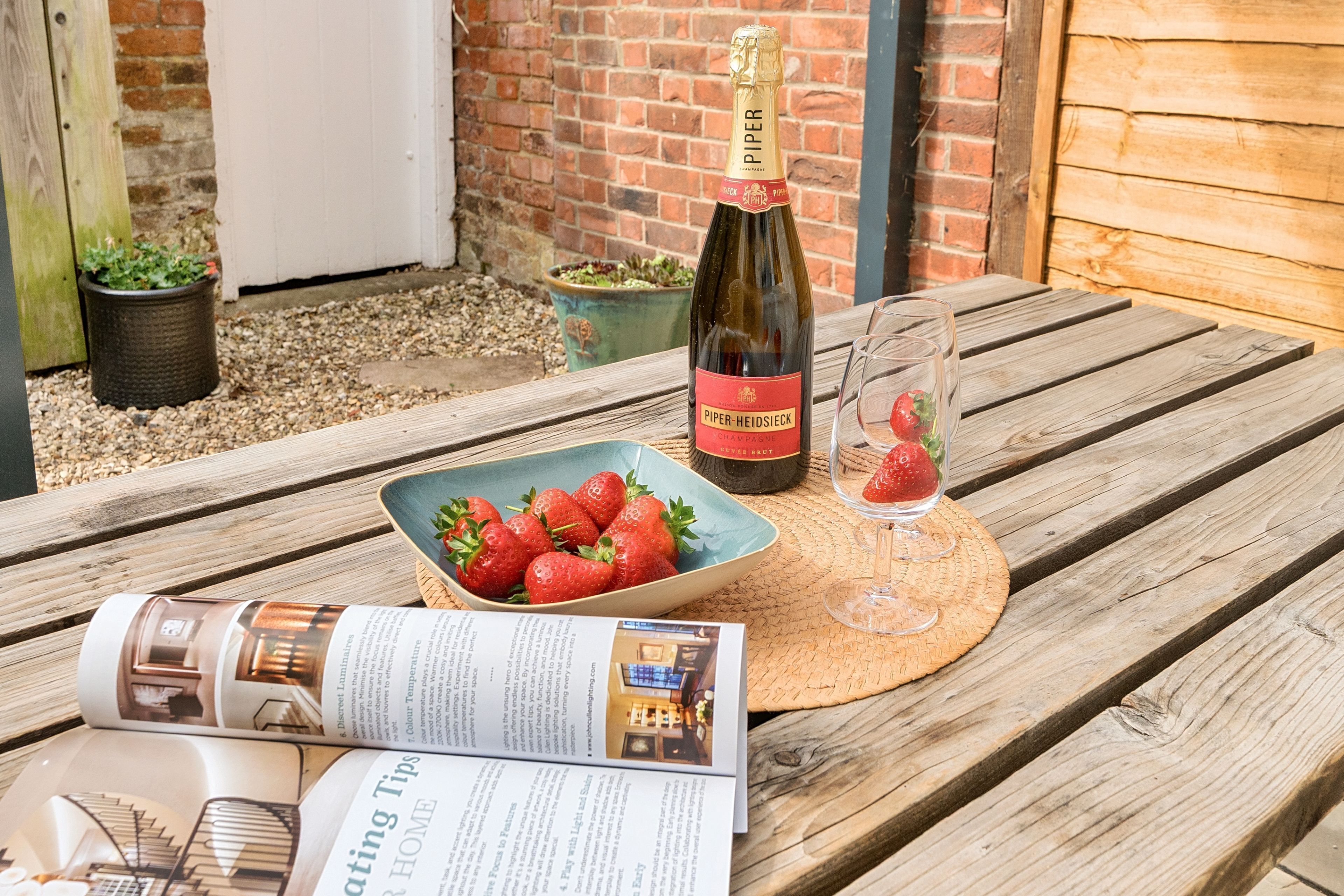 Ein Holztisch mit einer Flasche Piper Heidsieck Champagner, einer Schale Erdbeeren, einem Glas Erdbeersaft und einer Zeitschrift.