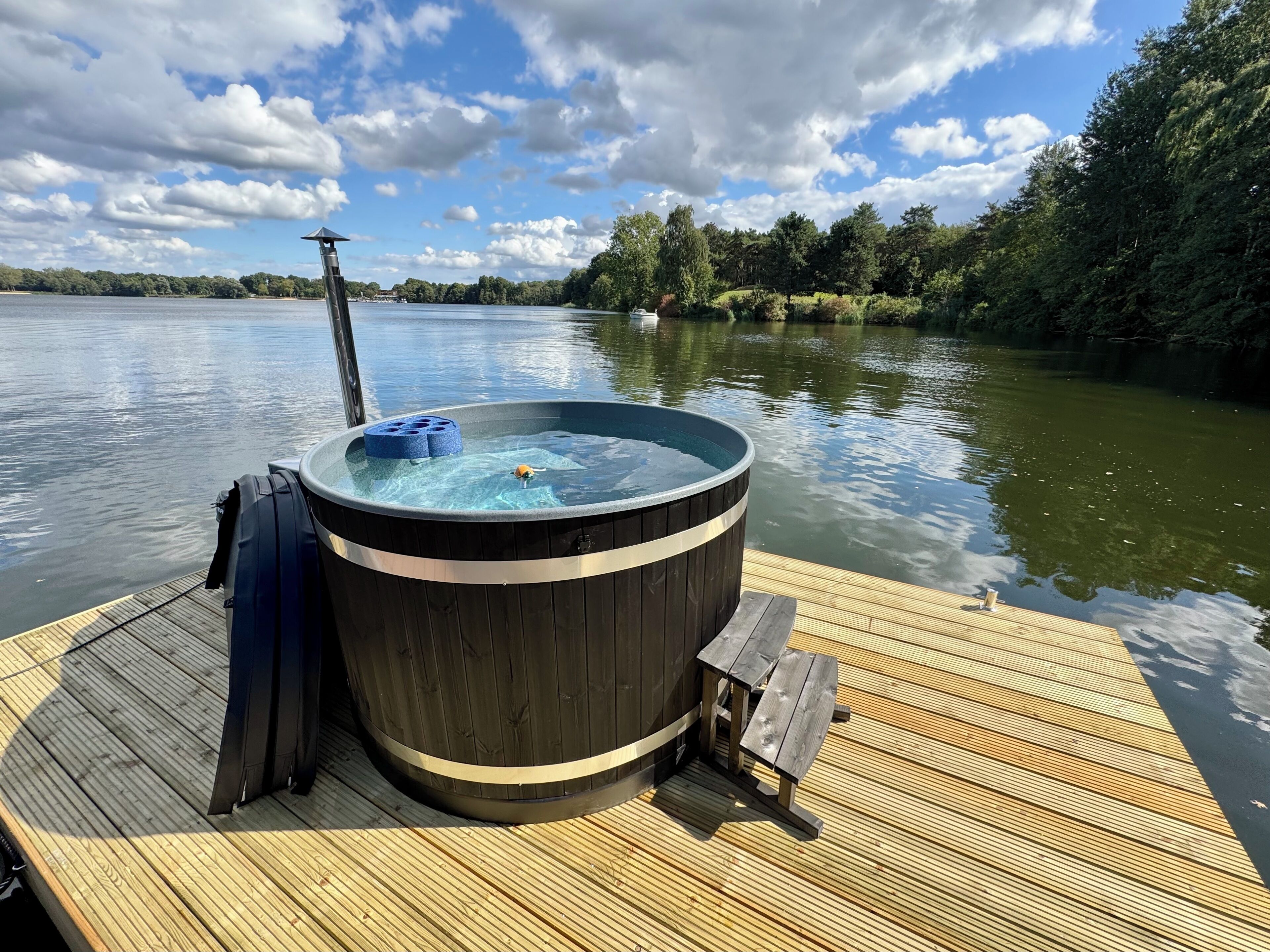 Een hottub op een houten terras aan een meer, met bomen op de achtergrond.