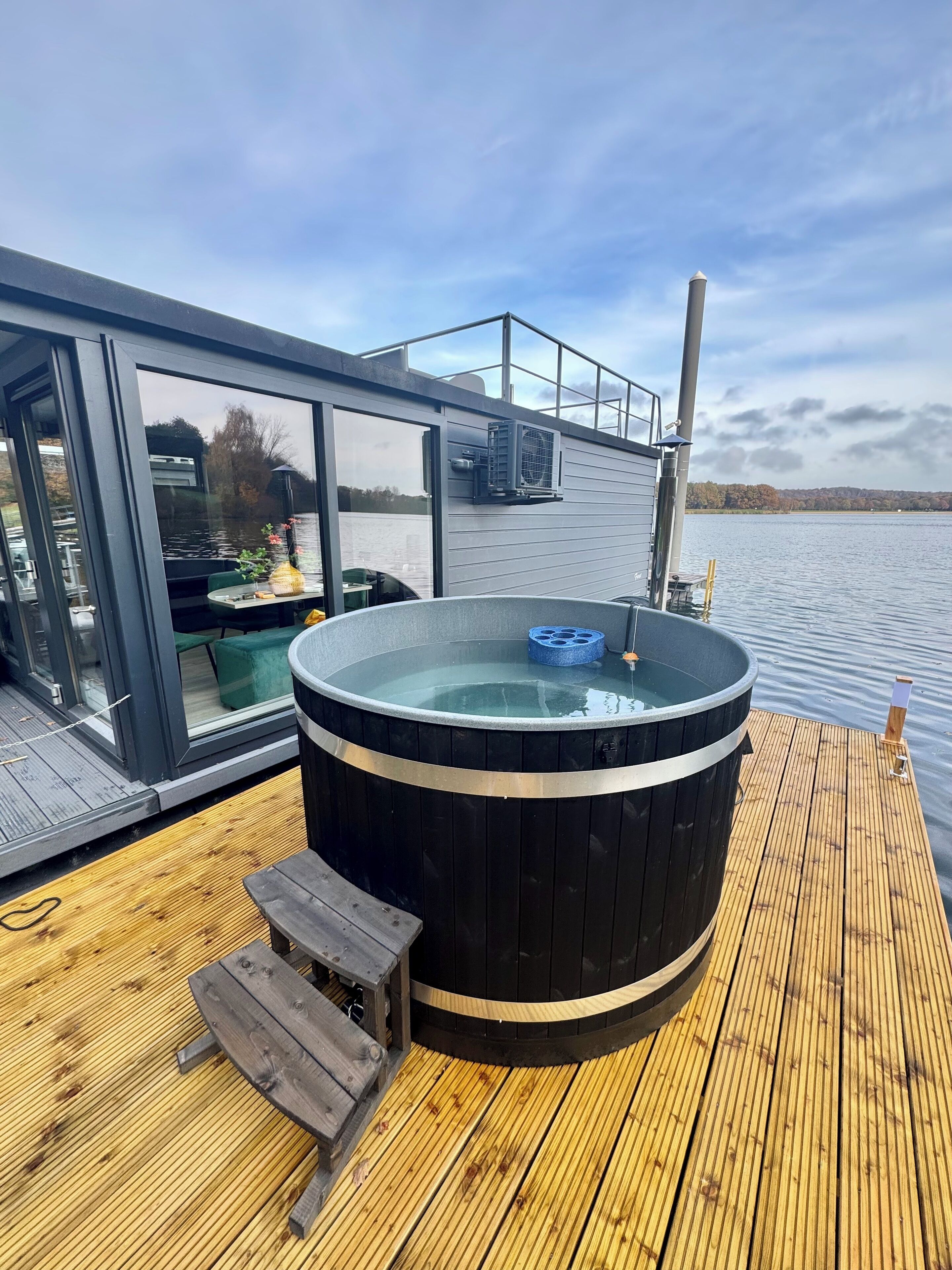 Un jacuzzi en una terraza de madera con cerramiento de vidrio y vista a un lago.