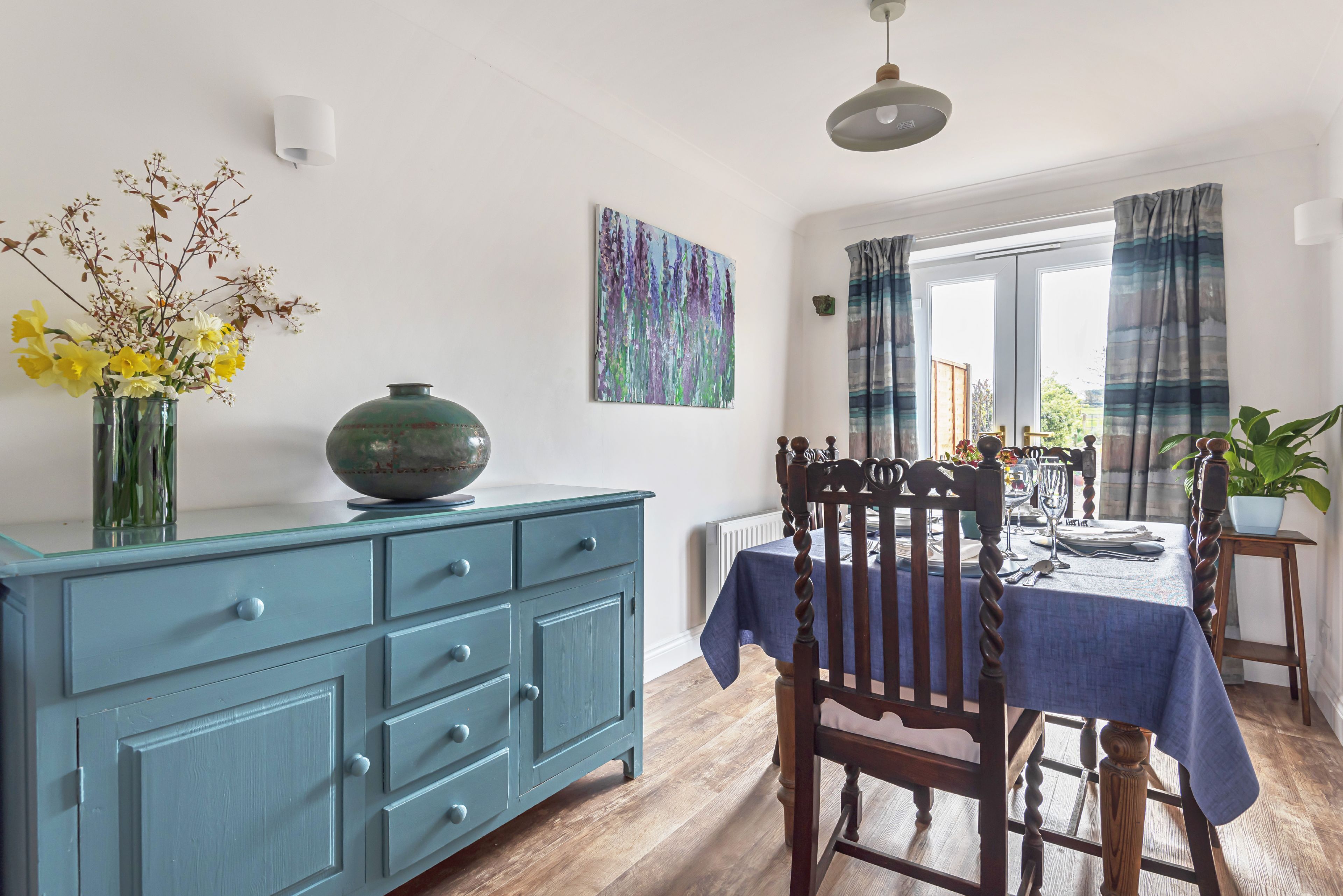 A dining area with a blue table, wooden chairs, a blue cabinet, and a vase with flowers.