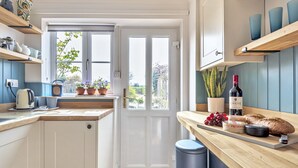 A kitchen with white cabinets, a wooden countertop, and a door leading outside. There are potted plants, a wine bottle, and a loaf of bread on the counter. - 4 Hare Cottage - sleeps 4 guests  in 3 bedrooms (South Creake)
