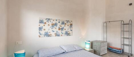 Un dormitorio con una cama, una obra de arte en la pared, una mesita de noche con lámpara, un armario y una estantería metálica.
