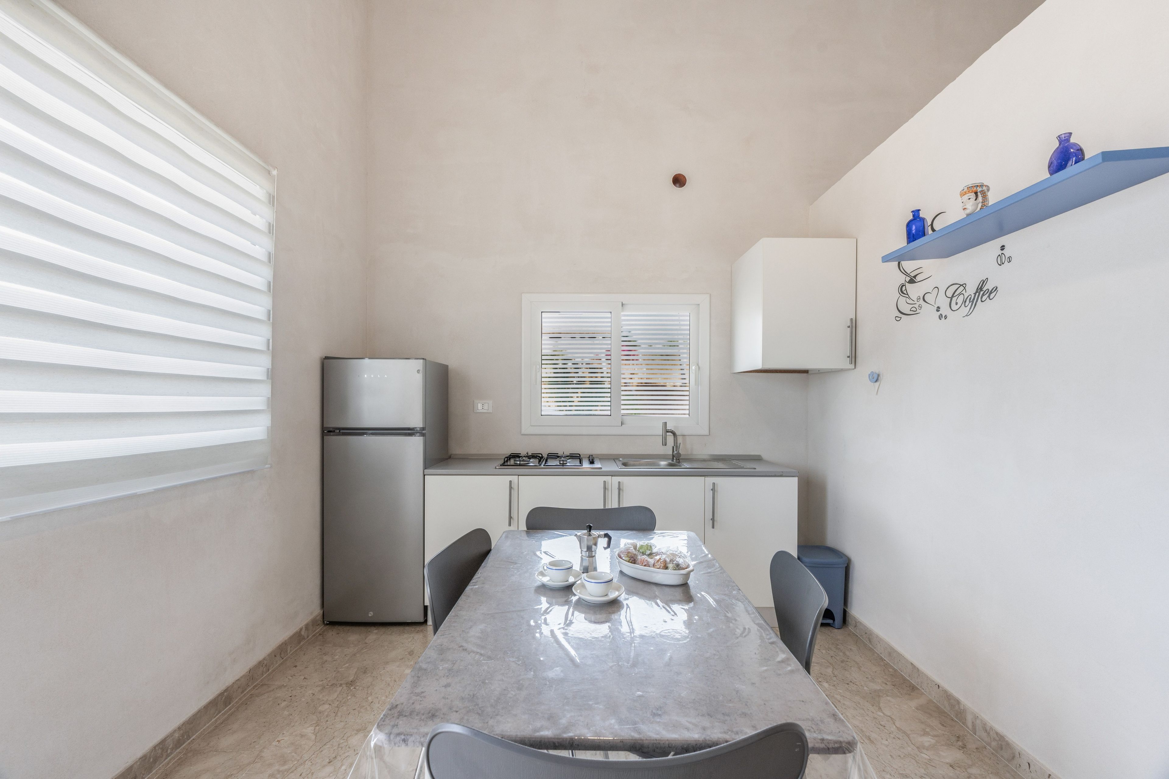 Une cuisine compacte équipée d’un réfrigérateur, d’une cuisinière et d’un évier. On y trouve une table à manger avec des chaises et une fenêtre avec des stores.