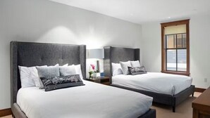 Hotellihuone, jossa on kaksi sänkyä, yöpöytä lampun kanssa ja ikkuna, jossa on sälekaihtimet.