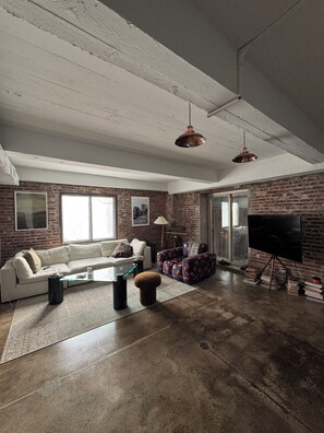 A spacious living room with a brick wall, a large flat-screen TV, a white sofa, and a coffee table. - Spacious loft style 2 bedroom 2 bath (Brooklyn)
