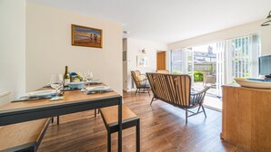 A dining area with a wooden table set for two, a chair with a cushioned backrest, and a view of the outdoors through a glass door.