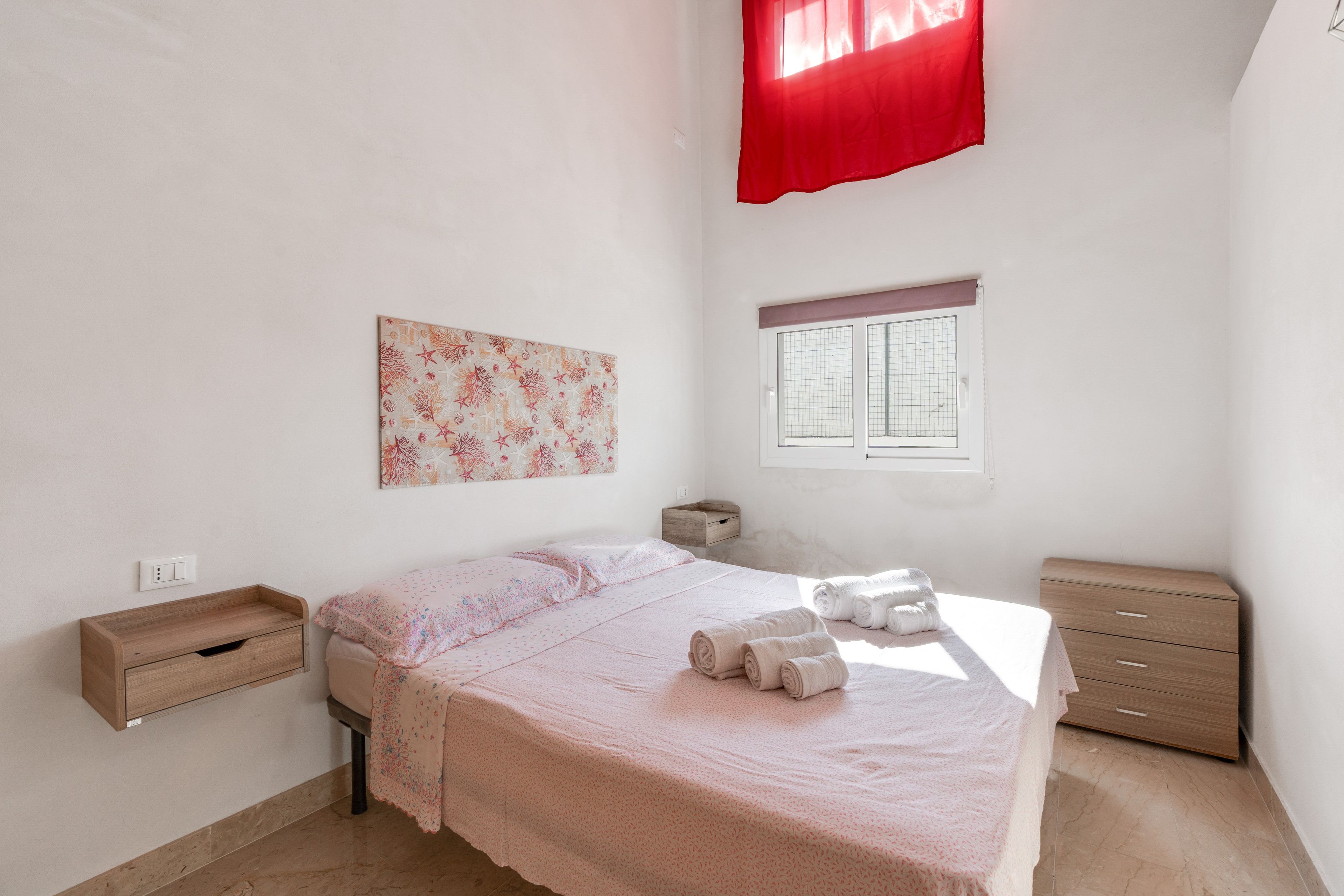 A bedroom with a bed, a bedside table, a window with a curtain, and a wall-mounted artwork.