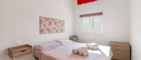 A bedroom with a bed, a bedside table, a window with a curtain, and a wall-mounted artwork.