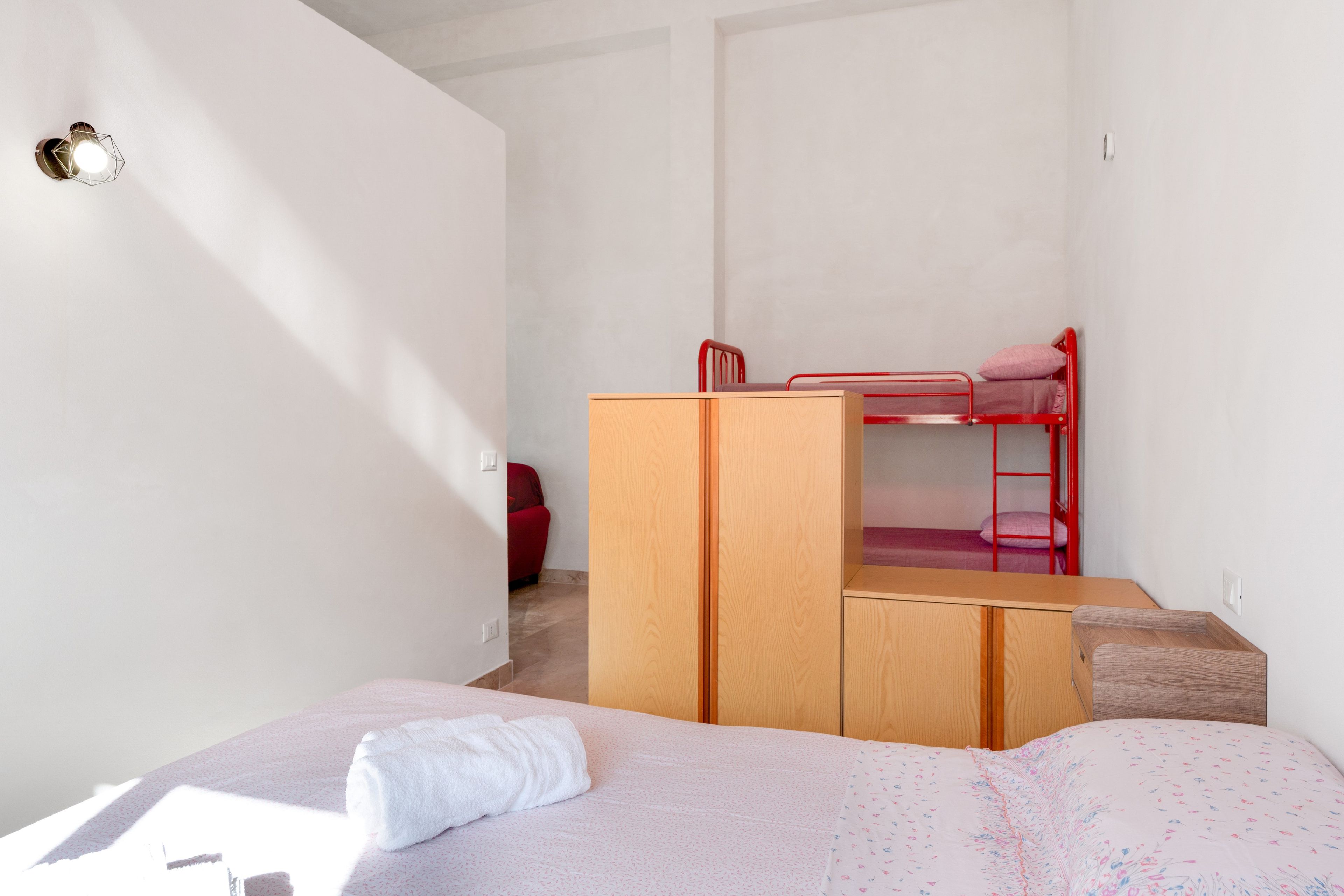Une chambre à coucher de taille réduite, avec un lit superposé, une armoire en bois et un lit simple sur lequel est posée une serviette enroulée.