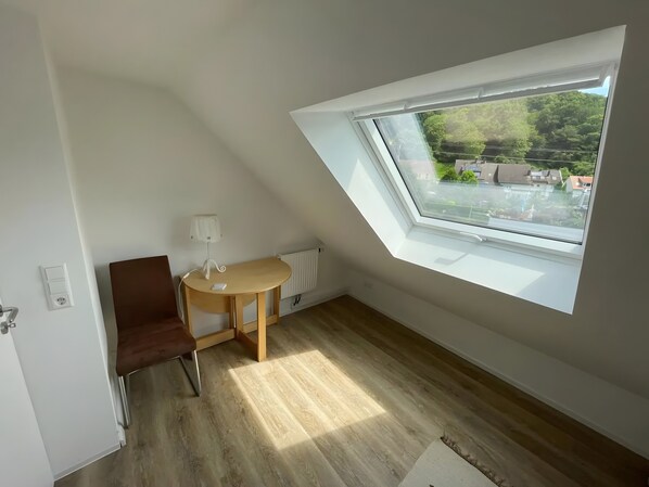 Une pièce avec un plafond en pente, une petite table avec une lampe, une chaise et une vue sur le ciel grâce à une lucarne.