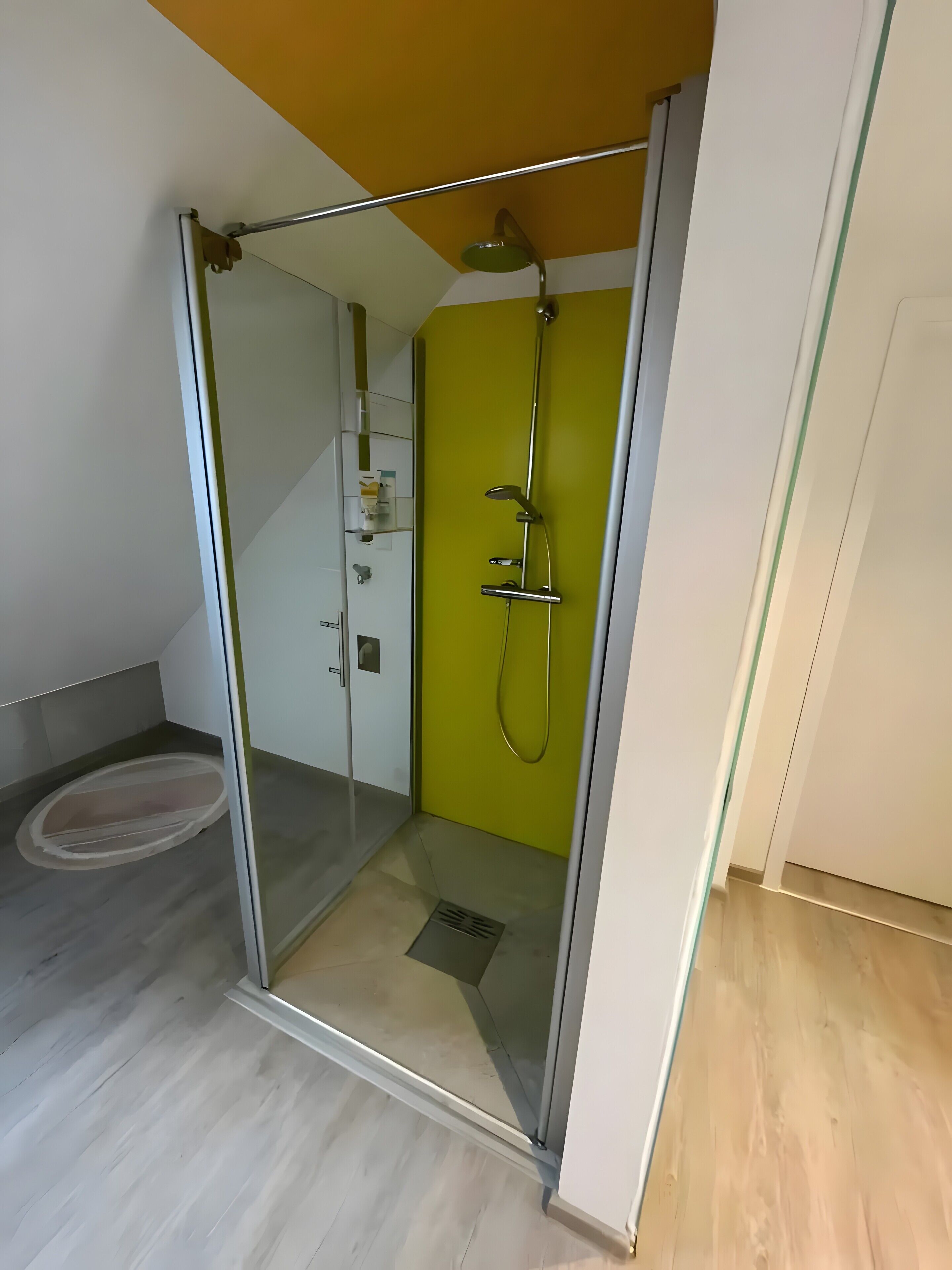 Eine begehbare, gläserne Dusche mit Duschbrause, einer weißen Tür und einer gelben Wand.