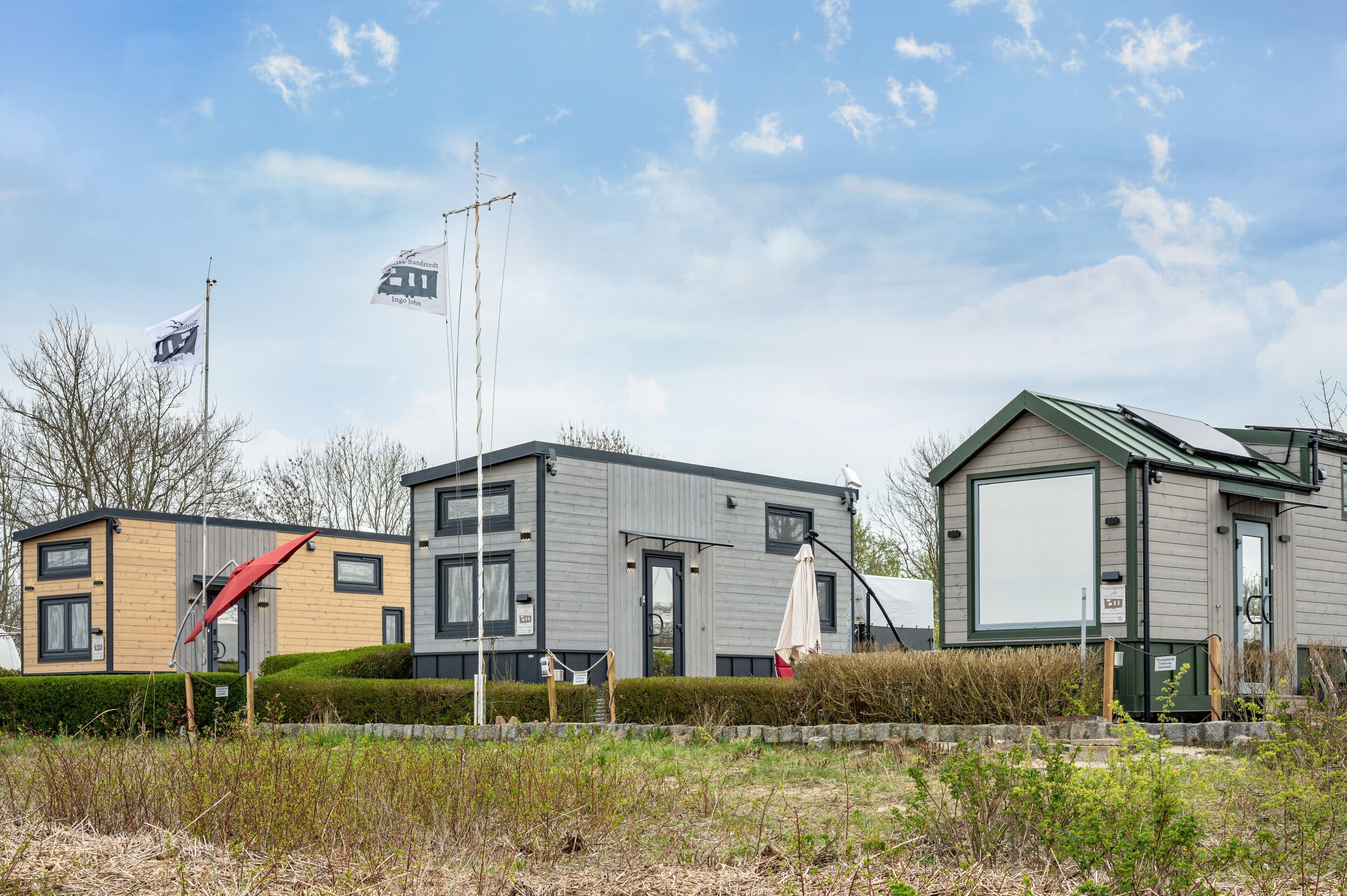A row of modern, single-story mobile homes with large windows and a flat roof, surrounded by a grassy area and a clear sky.