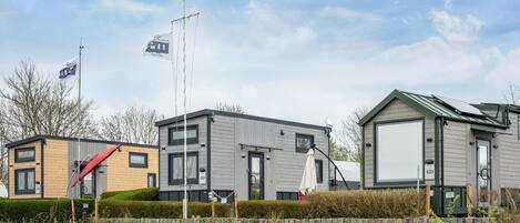 A row of modern, single-story mobile homes with large windows and a flat roof, surrounded by a grassy area and a clear sky.
