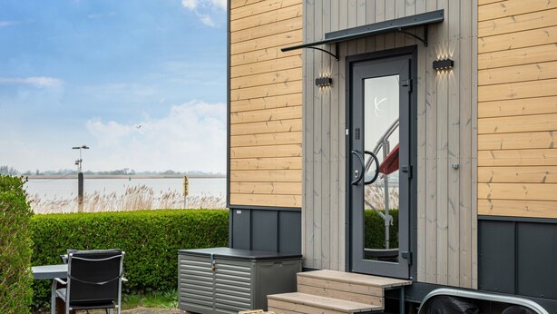 A modern building with a wooden exterior, a door with a glass panel, and a view of a body of water. - Tiny House 'Ingo John Vanessa' with Sea View, Wi-Fi, and Air Conditioning (Sandstedt)