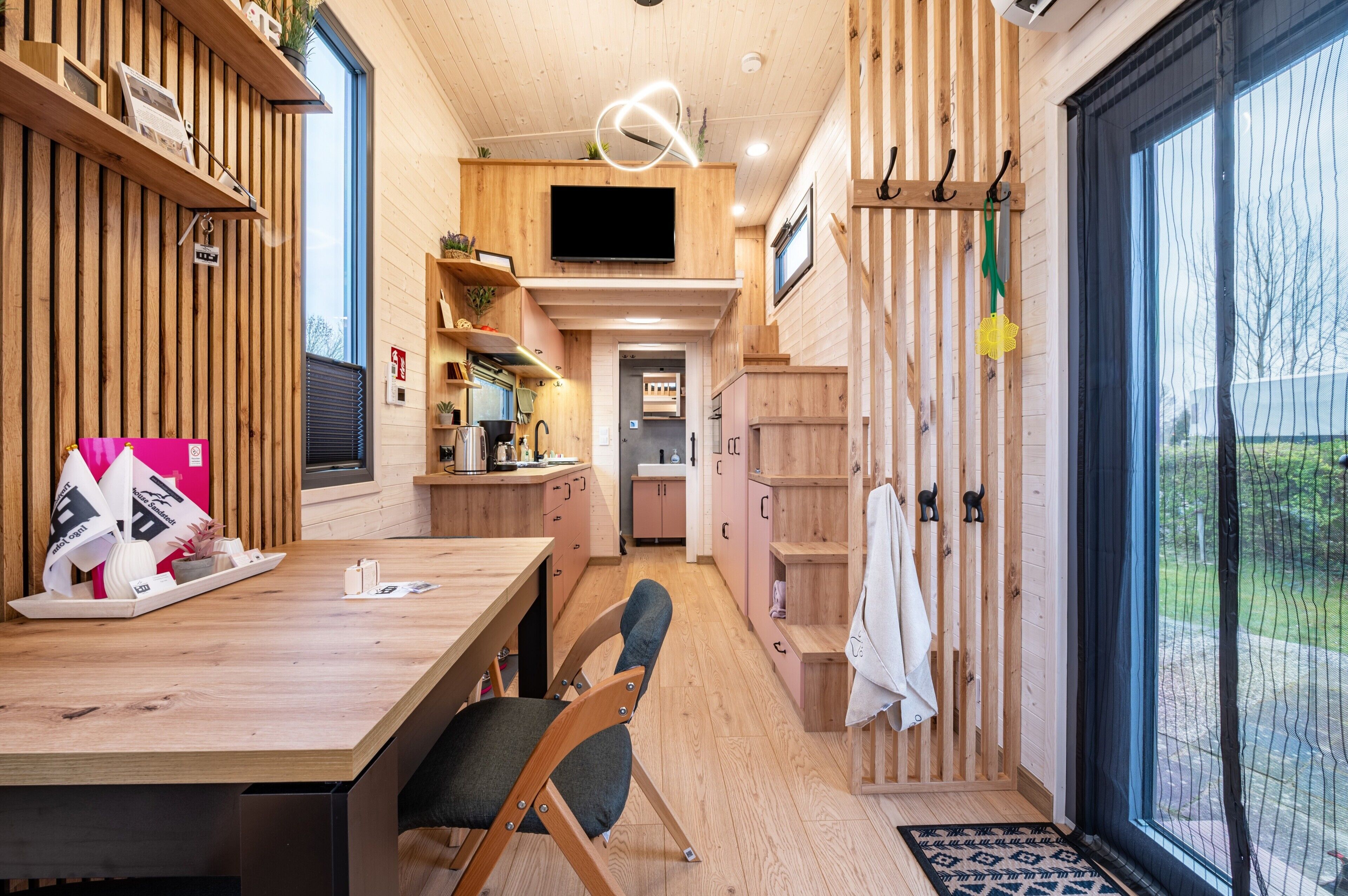 A modern interior with a wooden floor, a dining area with chairs, a kitchen with cabinets, a TV mounted on the wall, and a large window.