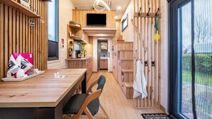 A modern interior with a wooden floor, a dining area with chairs, a kitchen with cabinets, a TV mounted on the wall, and a large window. - Tiny House 'Ingo John Vanessa' with Sea View, Wi-Fi, and Air Conditioning (Sandstedt)