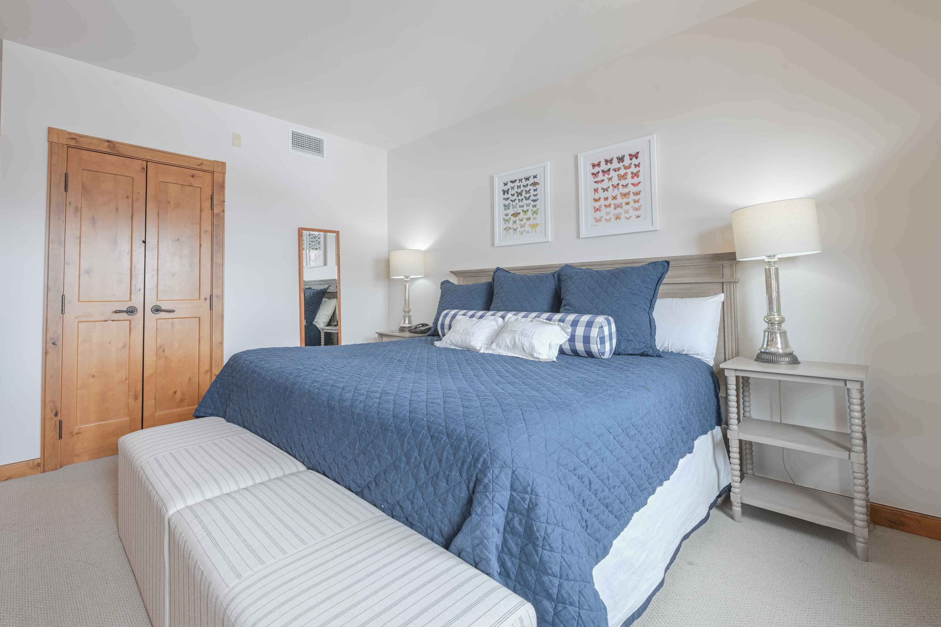 A bedroom with a large bed, a wooden wardrobe, a bedside table with a lamp, and a bench.