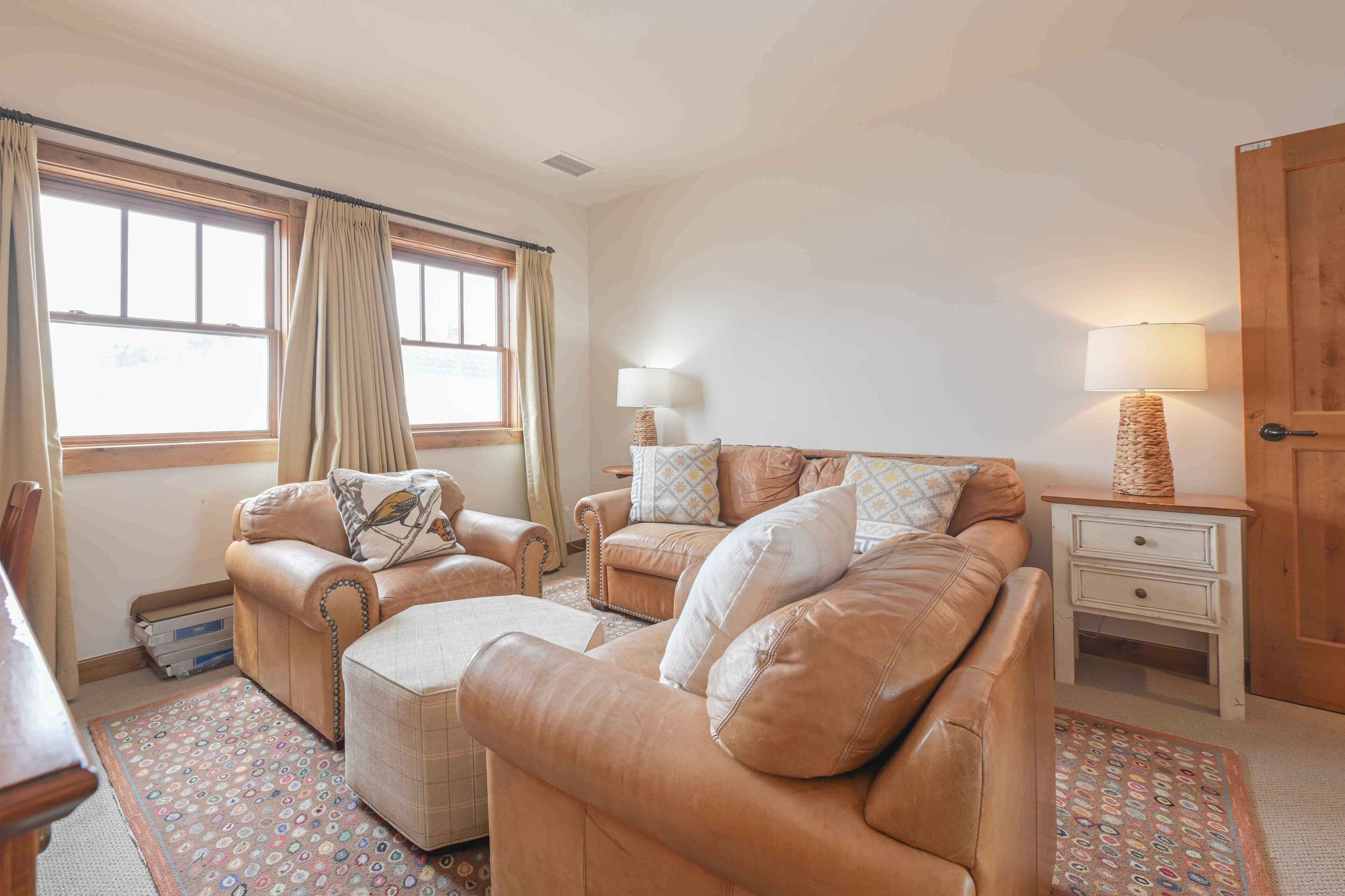A cozy living room with two leather sofas, a matching ottoman, a wooden side table with a lamp, and a patterned rug.