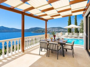 Eine Terrasse mit Pool, Esstisch und Stühlen, mit Blick auf eine malerische Landschaft aus Meer und Hügeln.