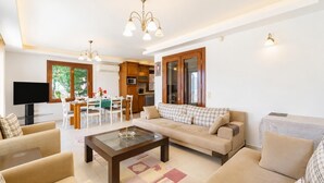 Un soggiorno moderno con divano, tavolino, TV, zona pranzo, cucina e ampie finestre.