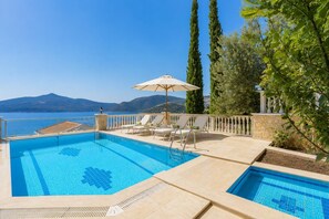 Ein Pool mit Liegestühlen und Sonnenschirm, umgeben von Grün und Blick aufs Meer.