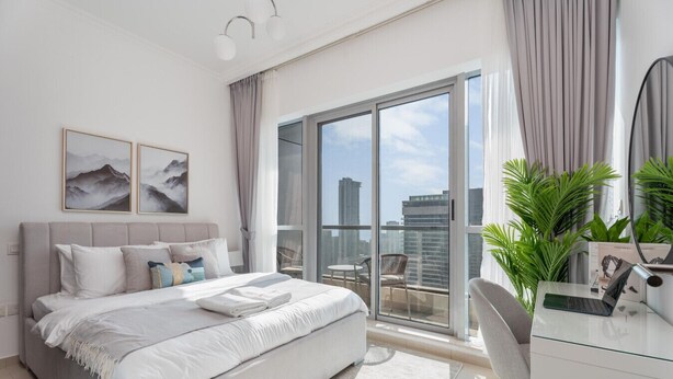 Un dormitorio moderno con una cama grande, un escritorio con una computadora portátil, un balcón con vistas a la ciudad y dos cuadros enmarcados en la pared.