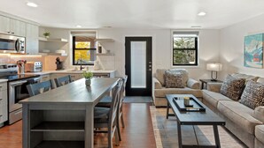 Una cocina moderna con electrodomésticos integrados, un comedor con mesa y sillas de madera, una sala de estar con sofá y una mesa de centro, y un ventanal que proporciona luz natural.