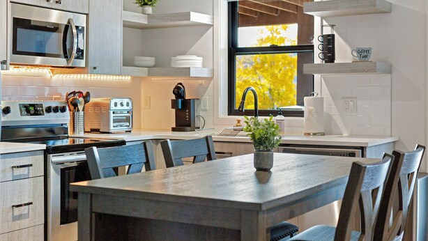 Una cocina moderna con horno empotrado, microondas y cafetera, una mesa de comedor con sillas y una ventana con vistas a árboles.