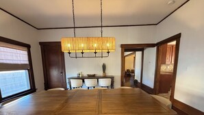 Ein Esszimmer mit einem großen Holztisch, vier weißen Stühlen, einer Pendelleuchte und Blick in die Küche durch einen offenen Durchgang.