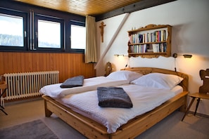 Uma cama de casal com estrutura de madeira, duas almofadas, uma mesa de cabeceira, uma estante, um radiador e uma janela com cortinas.