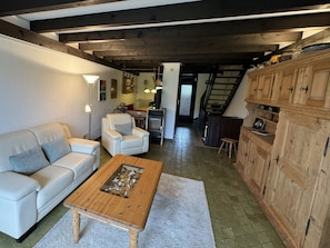 Sala de estar com sofá, poltrona, mesa de centro de madeira com conchas, lareira e área de cozinha com armários de madeira.