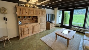 Habitació amb un armari de fusta, televisió, taula de dinar i vista cap a un paisatge verd a través de grans finestres.