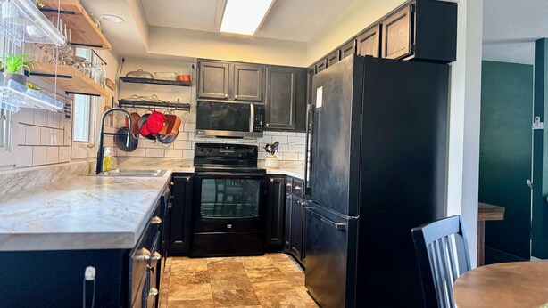 Cozinha moderna com armários escuros, geladeira preta, micro-ondas e forno. Há uma janela acima da pia, e uma planta é visível no balcão.