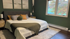 Quarto com uma cama grande, mesa de cabeceira, um detalhe de macramé na parede e janela com vista para árvores.