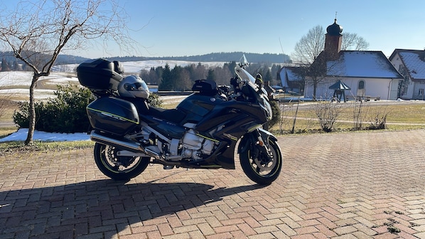 Une moto noire est garée sur une surface goudronnée, avec un paysage rural en arrière-plan.