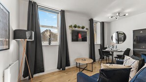 A modern living room with a sofa, a small round table, a TV, and a dining area with chairs. - Lovely Duplex for Long Stays - Parking (Dewsbury)