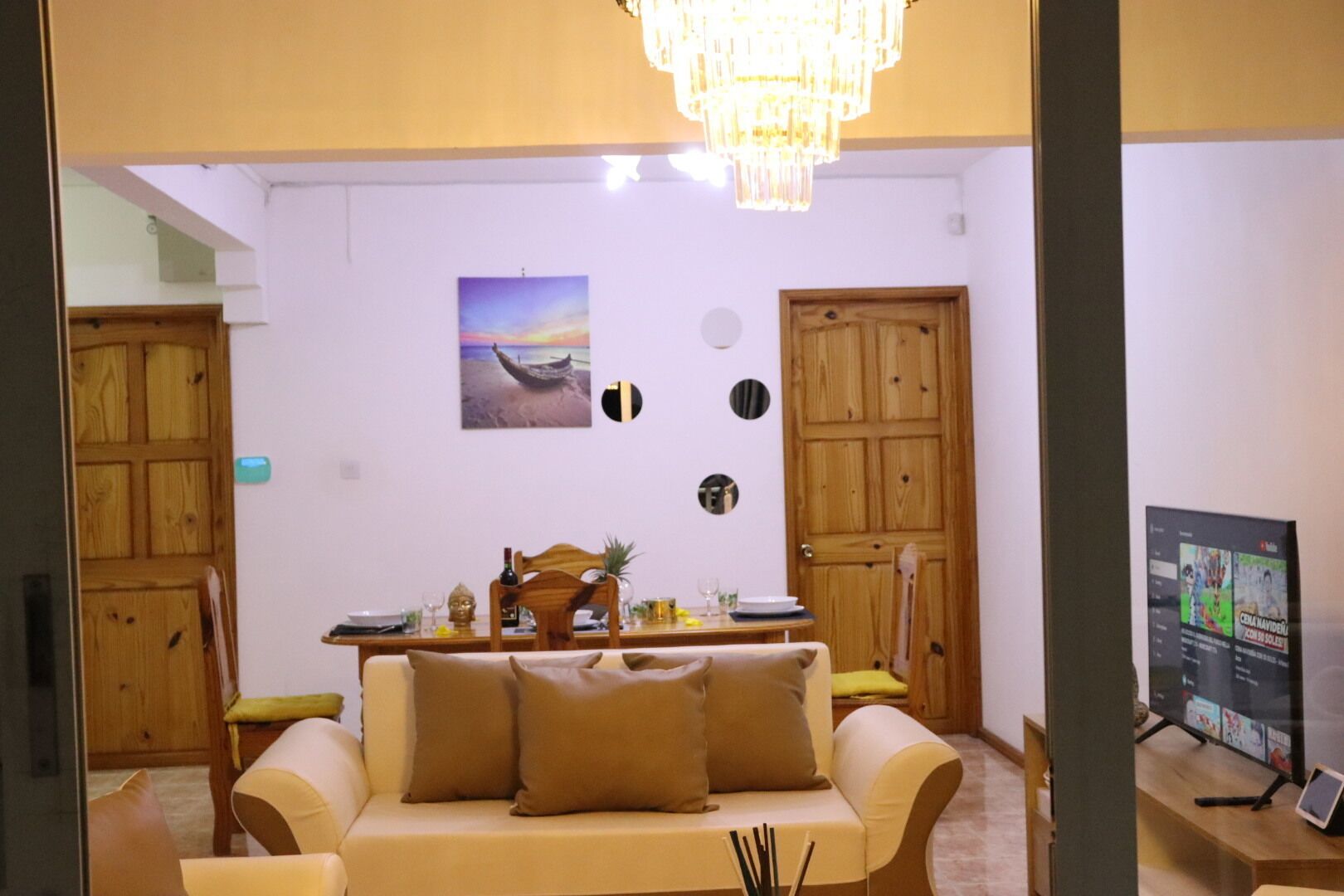 A living room with a sofa, a dining table, and a TV. There are wooden doors and a chandelier.