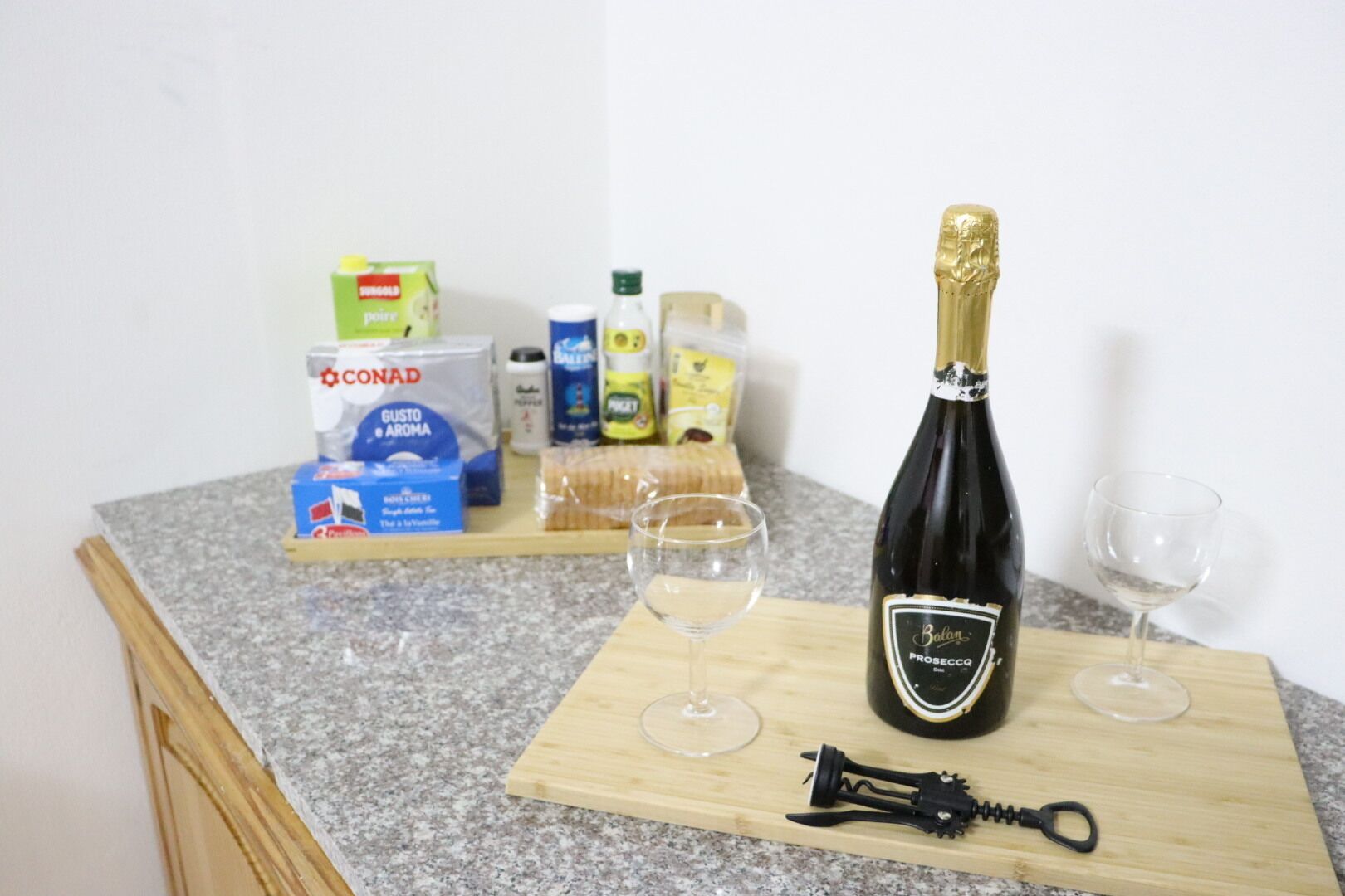 A kitchen counter with a bottle of Prosecco, two glasses, and various food items including a box of Conad pasta and a bottle of Ballantine olive oil.