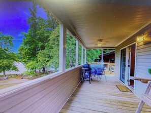 A covered porch with a grill, outdoor seating, and a view of trees and a building in the background. - Lakeside 2-bedroom house in adorable Delta with cool AC and charm (Delta)