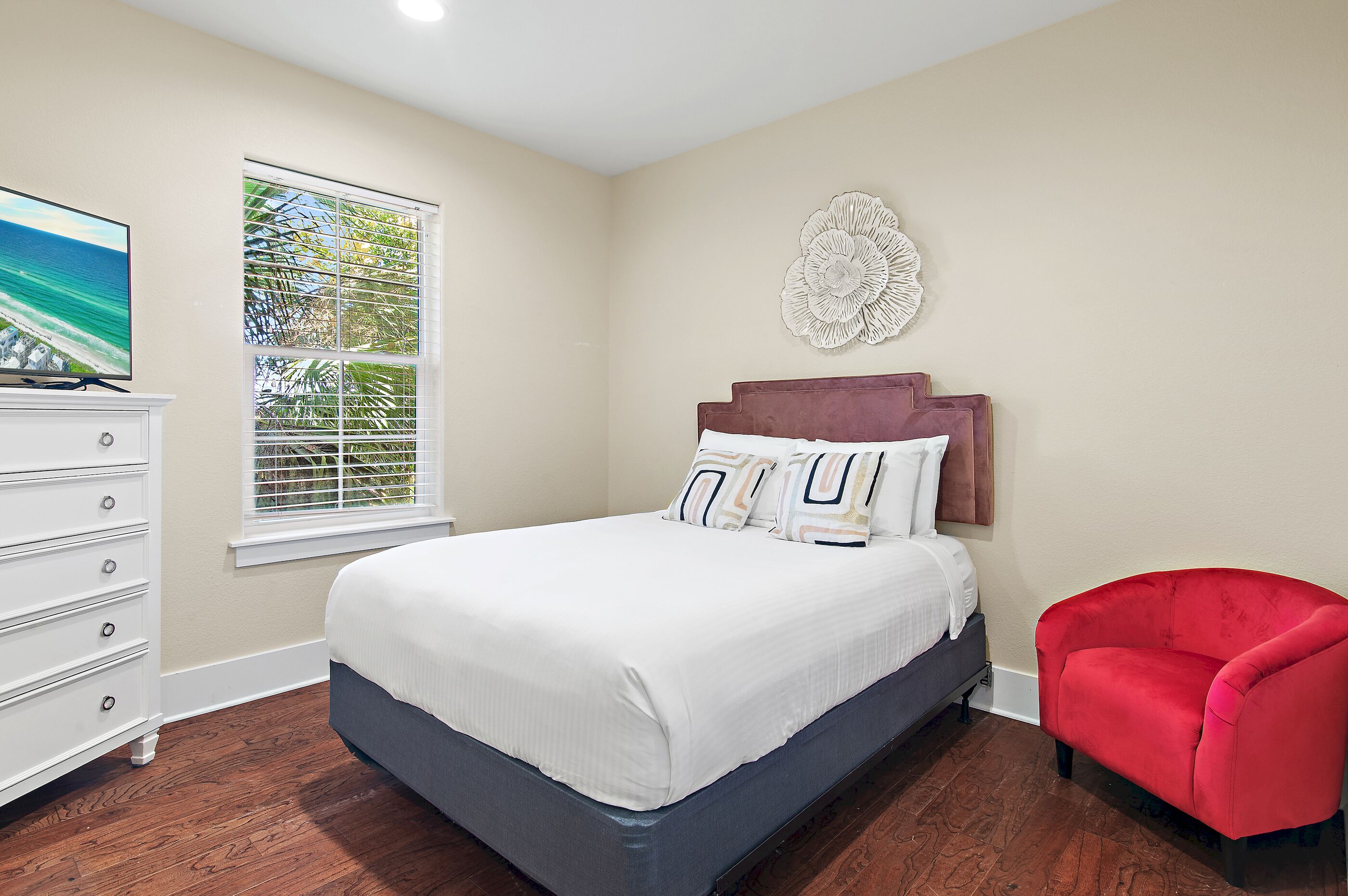 A bedroom with a bed, a dresser, a window with blinds, a red chair, and a wall decoration.