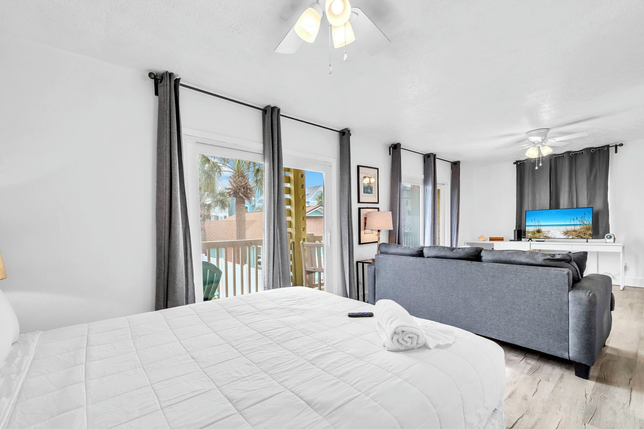 Quarto de hotel com cama, sofá cinza, televisão e varanda vista para palmeiras.